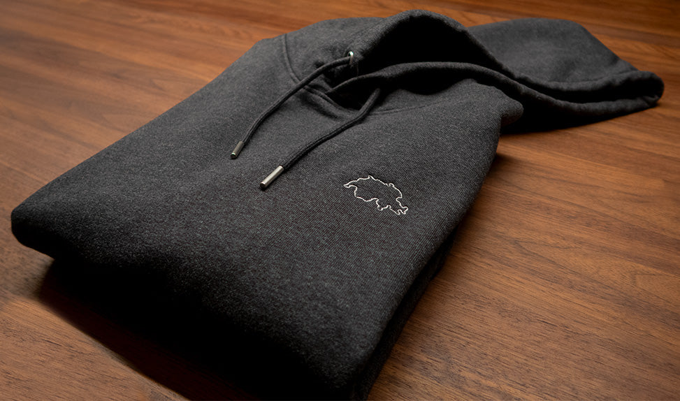 Dunkelgrauer unisex Twelvetia Pullover mit Kapuze und weiss-gestickter Schweizer Landesumrandung gefaltet auf holzfarbenen Tisch liegend