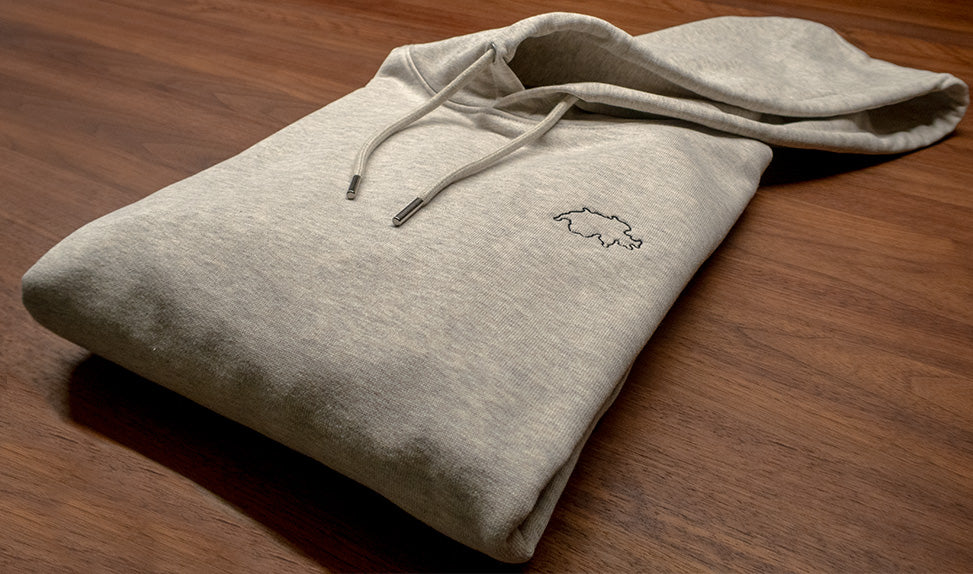 Grauer unisex Twelvetia Pullover mit Kapuze und schwarz-gestickter Schweizer Landesumrandung gefaltet auf holzfarbenen Tisch liegend