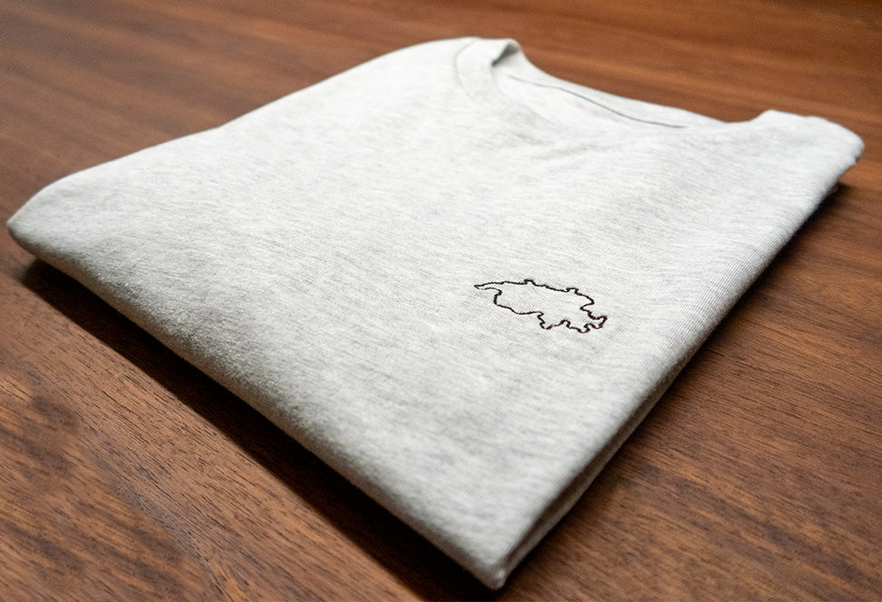 Graues unisex Twelvetia T-Shirt mit schwarz-gestickter Schweizer Landesumrandung gefaltet auf holzfarbenen Tisch liegend
