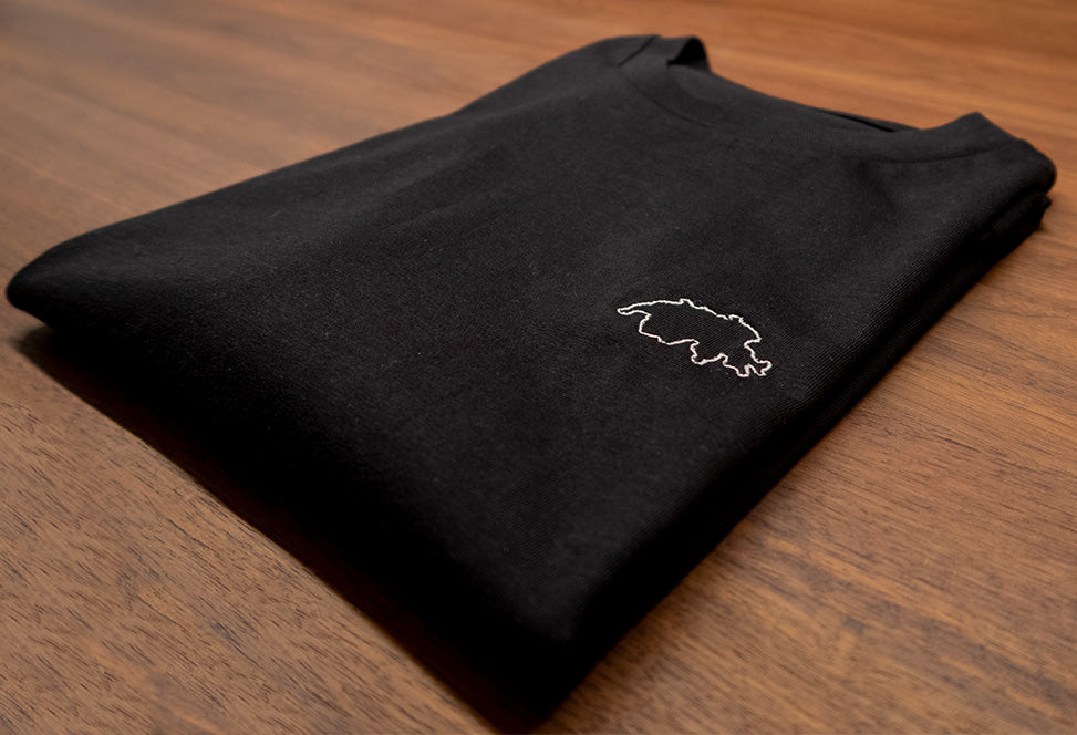 Schwarzes unisex Twelvetia T-Shirt mit weiss-gestickter Schweizer Landesumrandung gefaltet auf holzfarbenen Tisch liegend