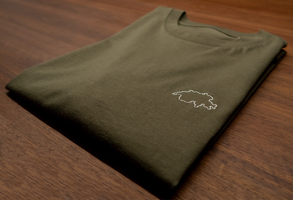 Khaki-farbenes unisex Twelvetia T-Shirt mit weiss-gestickter Schweizer Landesumrandung gefaltet auf holzfarbenen Tisch liegend