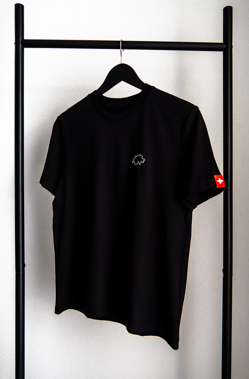 Schwarzes unisex Twelvetia T-Shirt mit weiss gestickter Schweizer Landesumrandung auf linker Brust und Schweizer-Flagge-Etikett am linken Ärmel. Hängend an schwarzem Bügel vor weisser Wand.