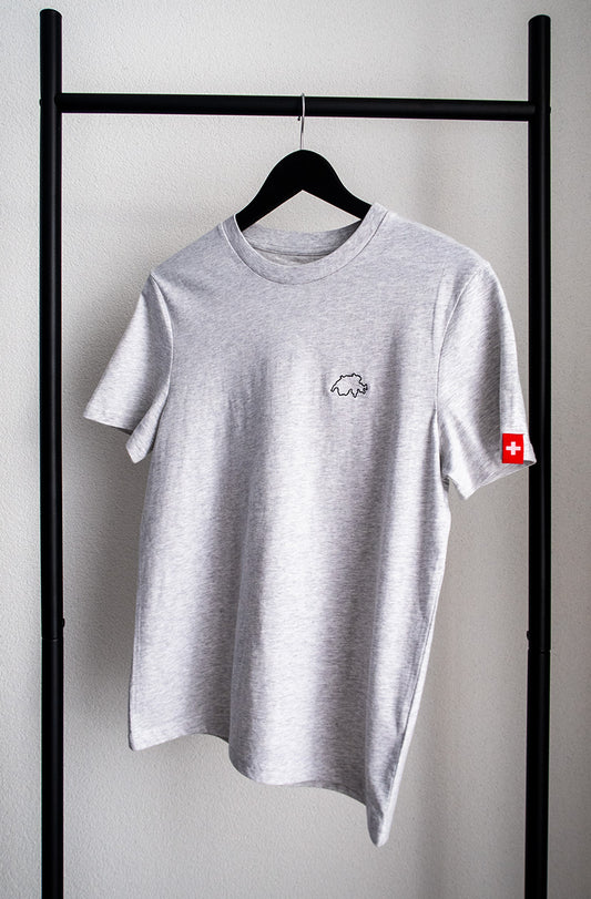 Graues unisex Twelvetia T-Shirt mit schwarz gestickter Schweizer Landesumrandung auf linker Brust und Schweizer-Flagge-Etikett am linken Ärmel. Hängend an schwarzem Bügel vor weisser Wand.
