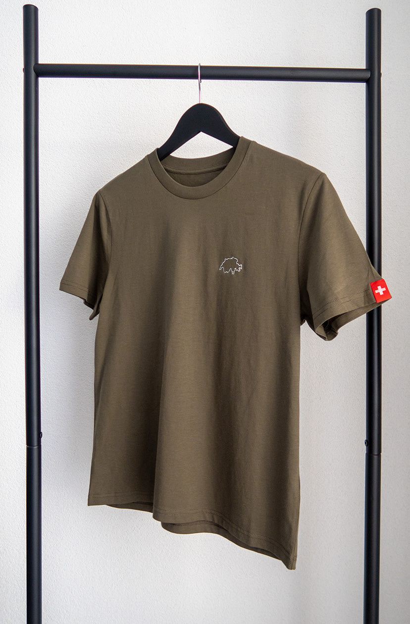 Khaki-farbenes unisex Twelvetia T-Shirt mit weiss gestickter Schweizer Landesumrandung auf linker Brust und Schweizer-Flagge-Etikett am linken Ärmel. Hängend an schwarzem Bügel vor weißer Wand.