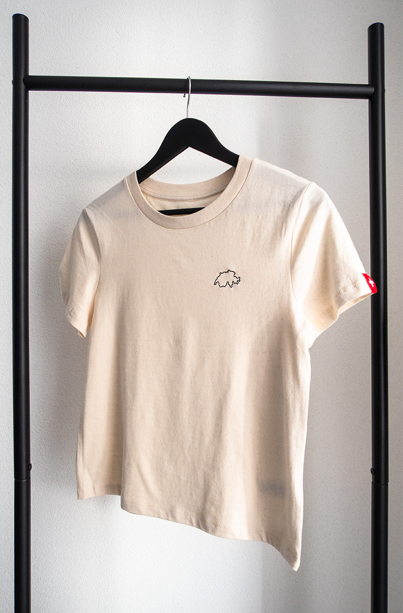 Beiges Twelvetia Damen-T-Shirt mit schwarz gestickter Schweizer Landesumrandung auf linker Brust und Schweizer-Flagge-Etikett am linken Ärmel. Hängend an schwarzem Bügel vor weißer Wand.
