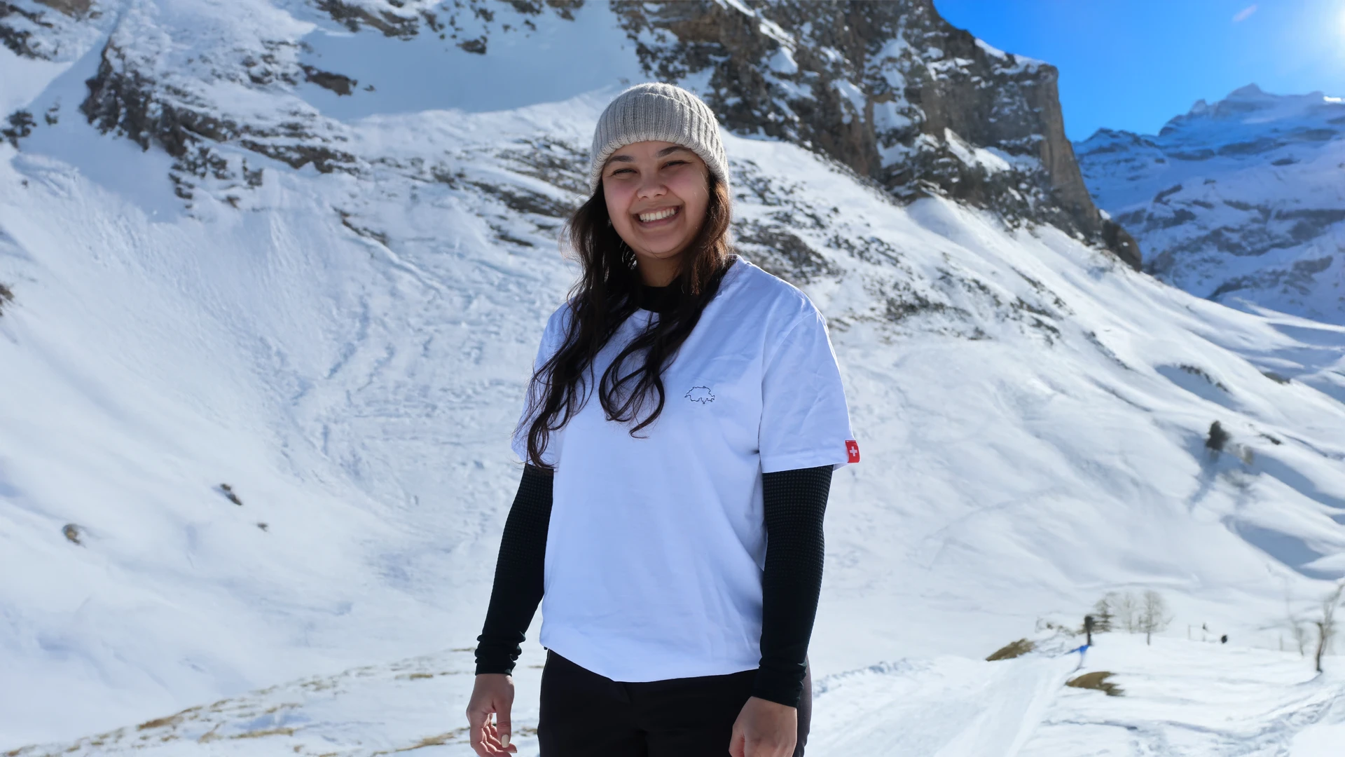 Ein weibliches Modell, welches ein weisses Twelvetia T-Shirt mit schwarz-gestickter Schweizer Landesumrandung und einem Ärmelettikett der Schweizer Fahne vor einer schneebedeckten Bergkulisse trägt.