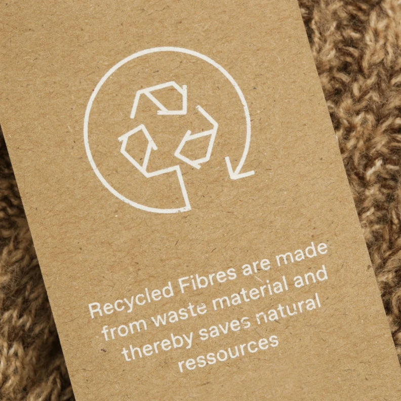 Ein Textilschildchen mit dem Recyclingsymbol und der Aufschrift „Recycled fibres are made from waste material and thereby saves natural ressources“ liegt auf braunem Stoff. Das Bild verdeutlicht Twelvetias Engagement für Recycling und die Verwendung umweltfreundlicher Materialien.