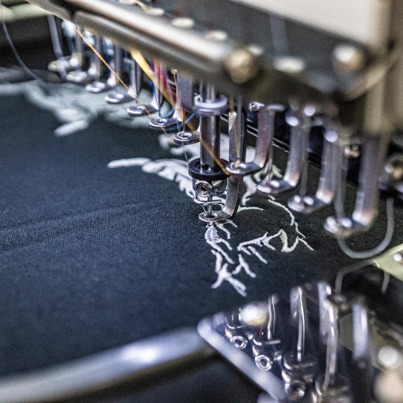 Eine industrielle Stickmaschine bestickt ein schwarzes Kleidungsstück mit einem Logo. Das Bild steht für die hochwertige Verarbeitung und die sorgfältige Veredelung der Textilien bei Twelvetia.